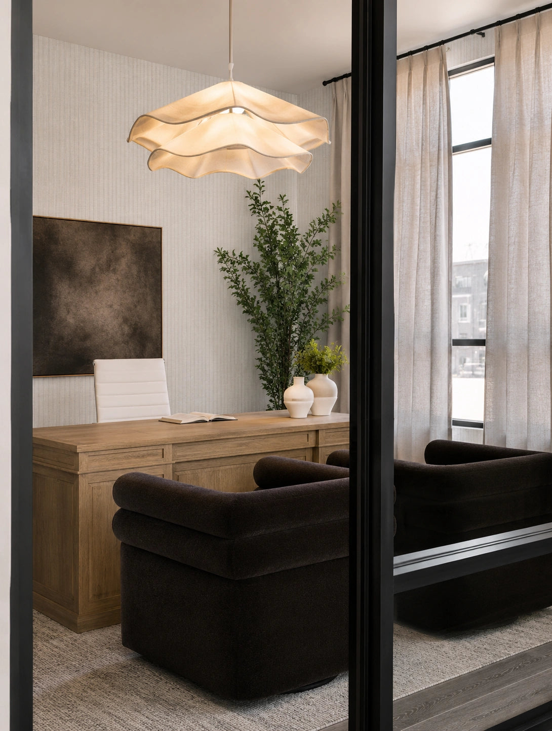 A modern home office interior featuring a light wood desk, a dark brown velvet armchair, a wavy beige pendant light, and a tall green plant against a textured wall.