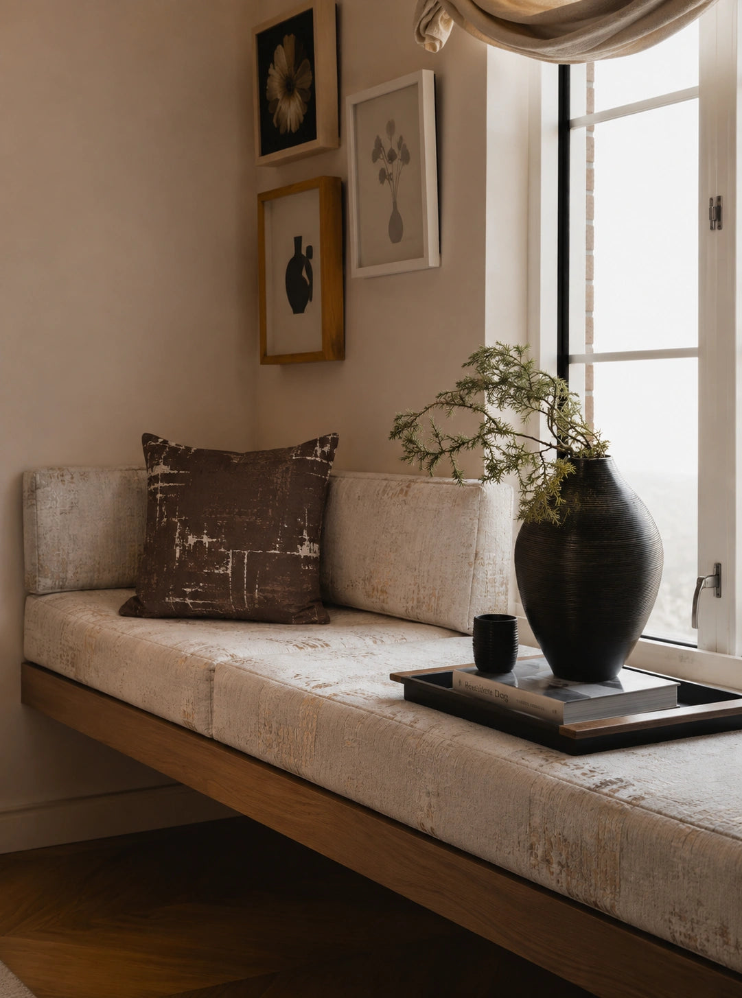 A modern floating daybed with textured cream upholstery and a dark brown accent pillow, styled in a bright room next to a window with a large black ceramic vase on a tray.