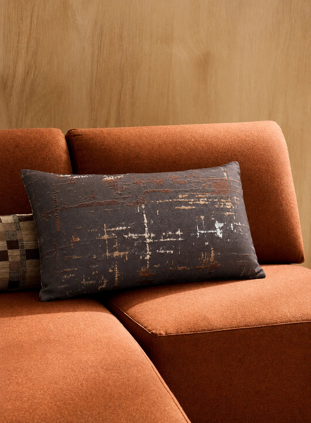 A rectangular throw pillow with a distressed dark gray and rust-orange abstract pattern, styled on a rust-orange sectional sofa against a light wood wall.
