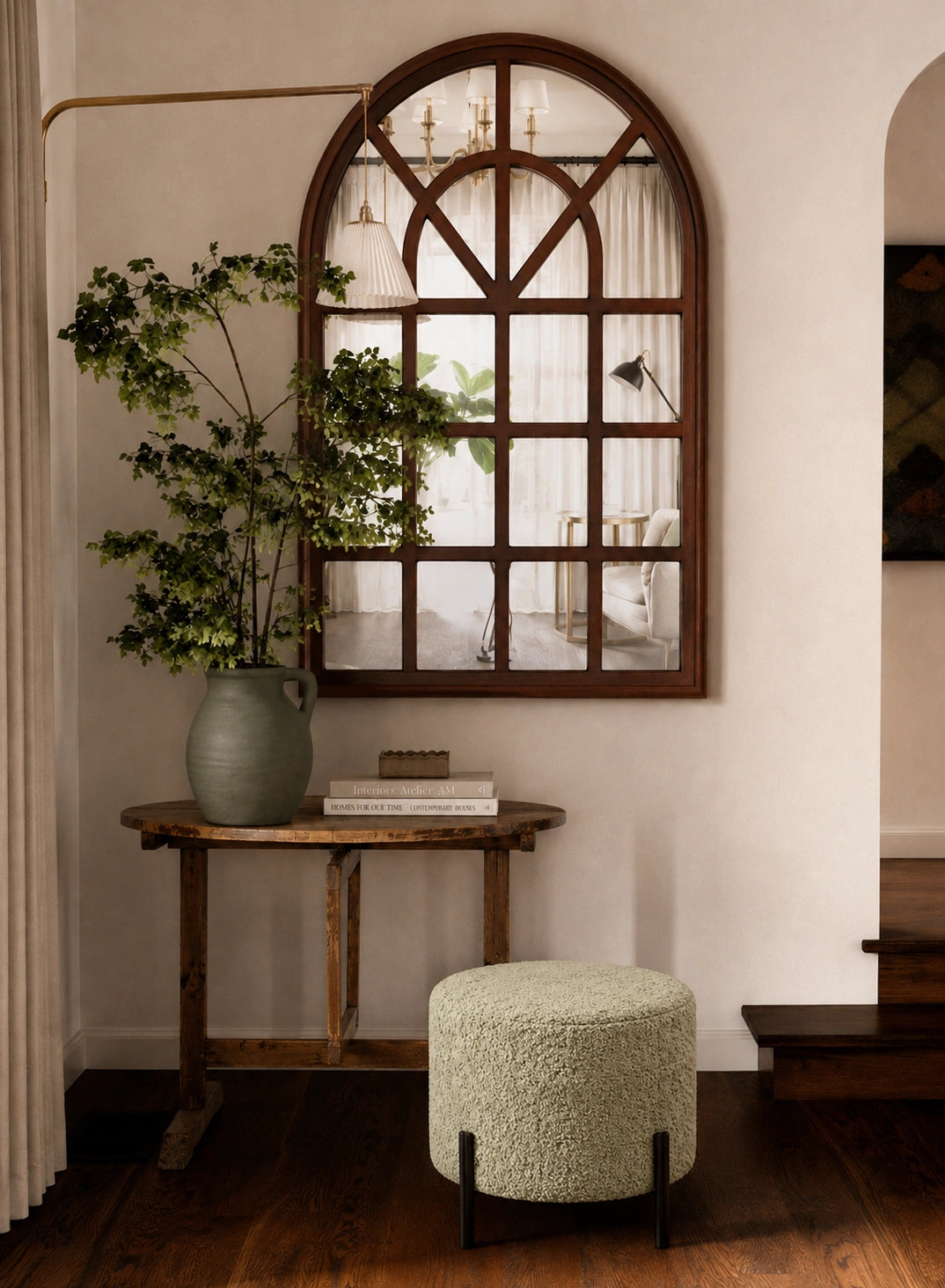 A styled interior corner featuring a large arched wood-framed mirror on a cream wall, a rustic wooden console table with a green ceramic planter holding a leafy plant, a round textured light green pouf, and dark wood flooring.