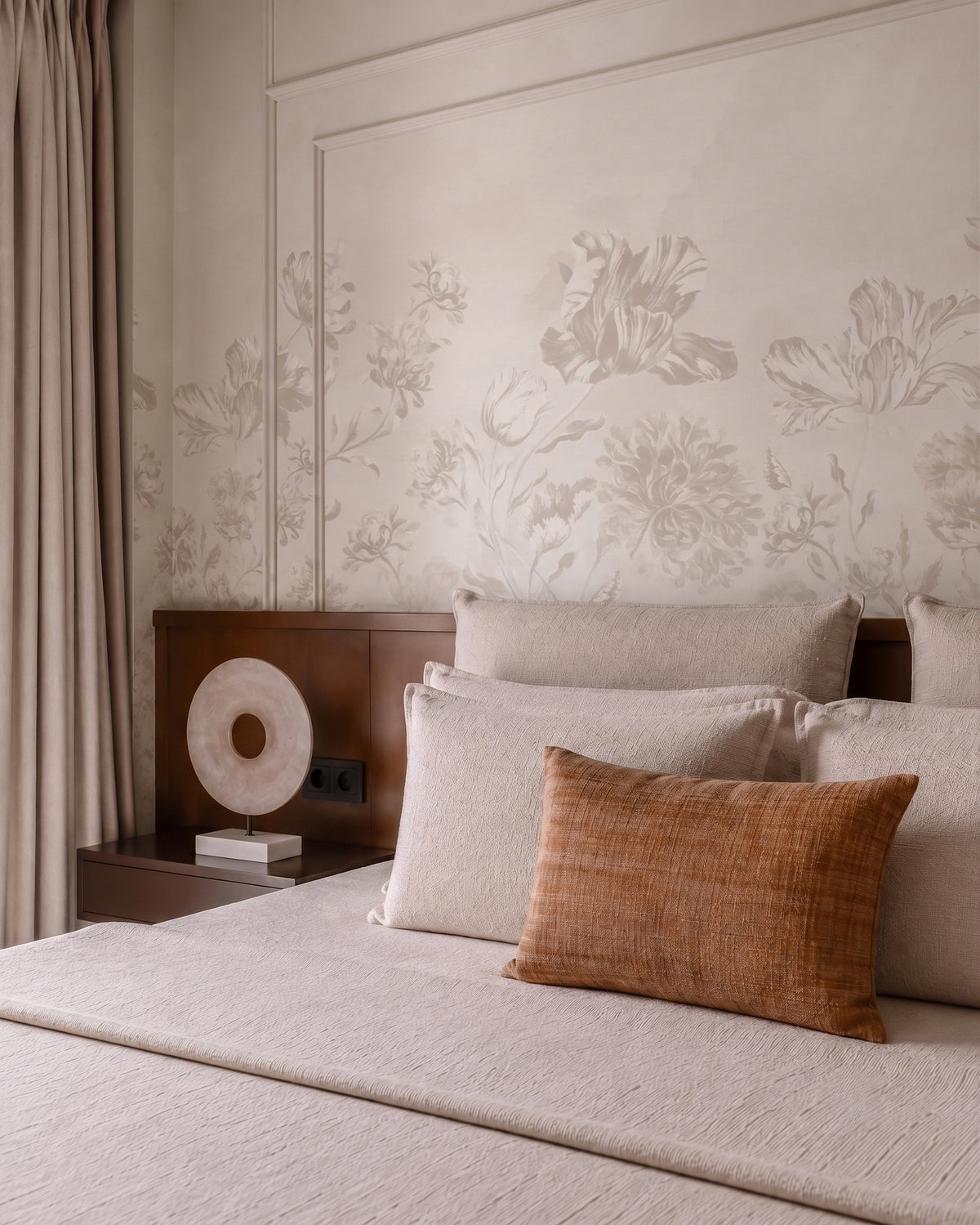 A styled bedroom scene featuring a bed with neutral beige bedding, a textured rust-orange accent pillow, a dark wood headboard, and a wall with matching beige floral wallpaper.