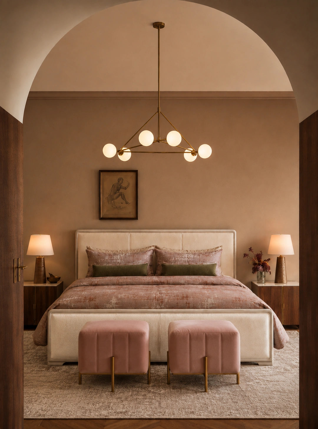 A styled bedroom interior viewed through an arched doorway, featuring a bed with pink bedding, two pink ottomans at the foot, bedside tables with lamps, and a modern multi-bulb chandelier hanging above.