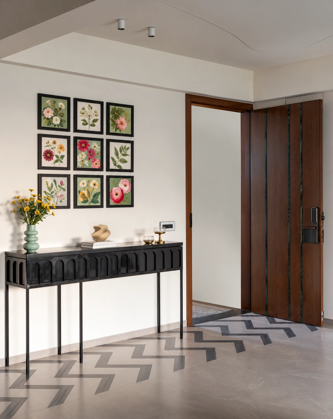 Modern entryway featuring a black console table, colorful botanical gallery wall, decorative vase and flowers, contemporary wood front door, and geometric floor detailing in a bright neutral interior.