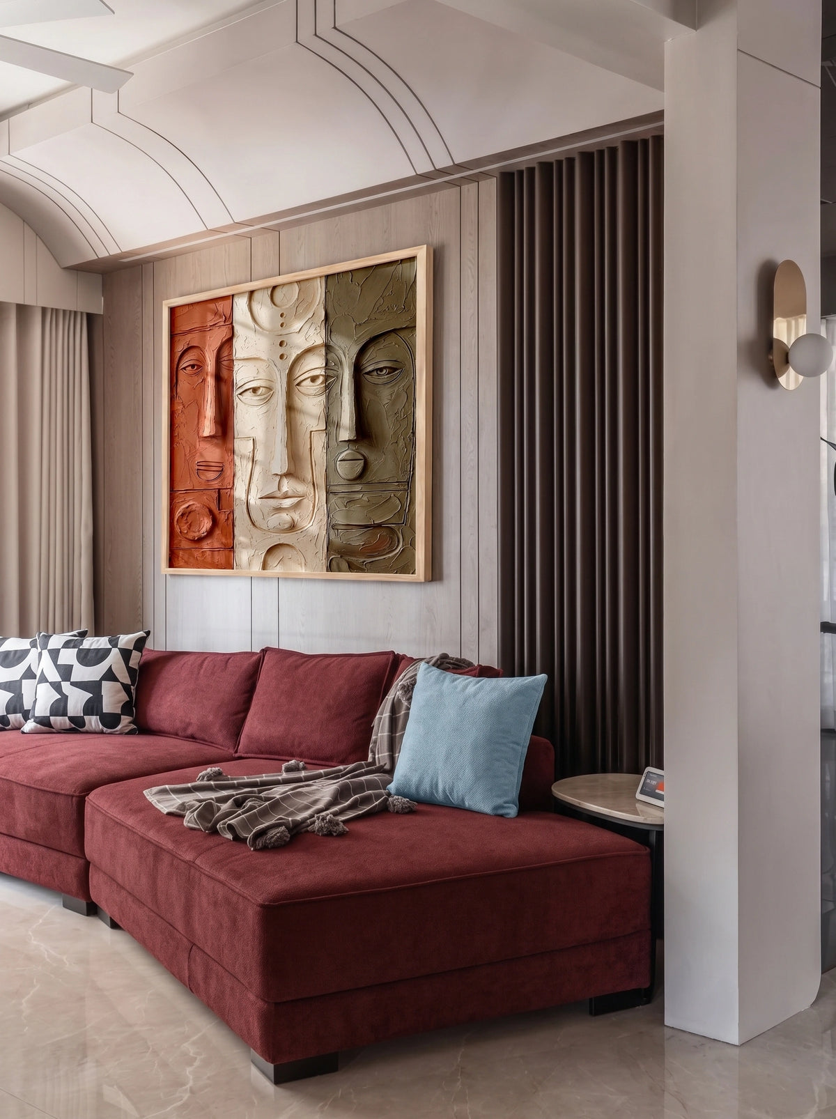 Modern living room with a deep burgundy sectional sofa, blue and patterned cushions, a textured three-face abstract artwork, warm wood wall paneling, dark vertical slats, and sculptural contemporary lighting.