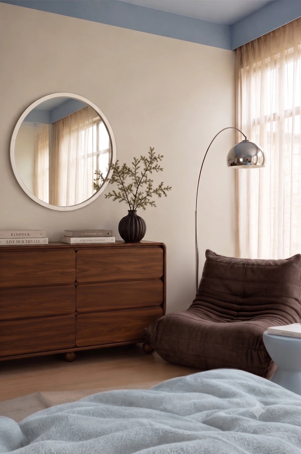 Cozy modern bedroom with a walnut wood dresser, round wall mirror, dark brown lounge chair, curved floor lamp, black decorative vase, sheer curtains, and soft blue ceiling accents.