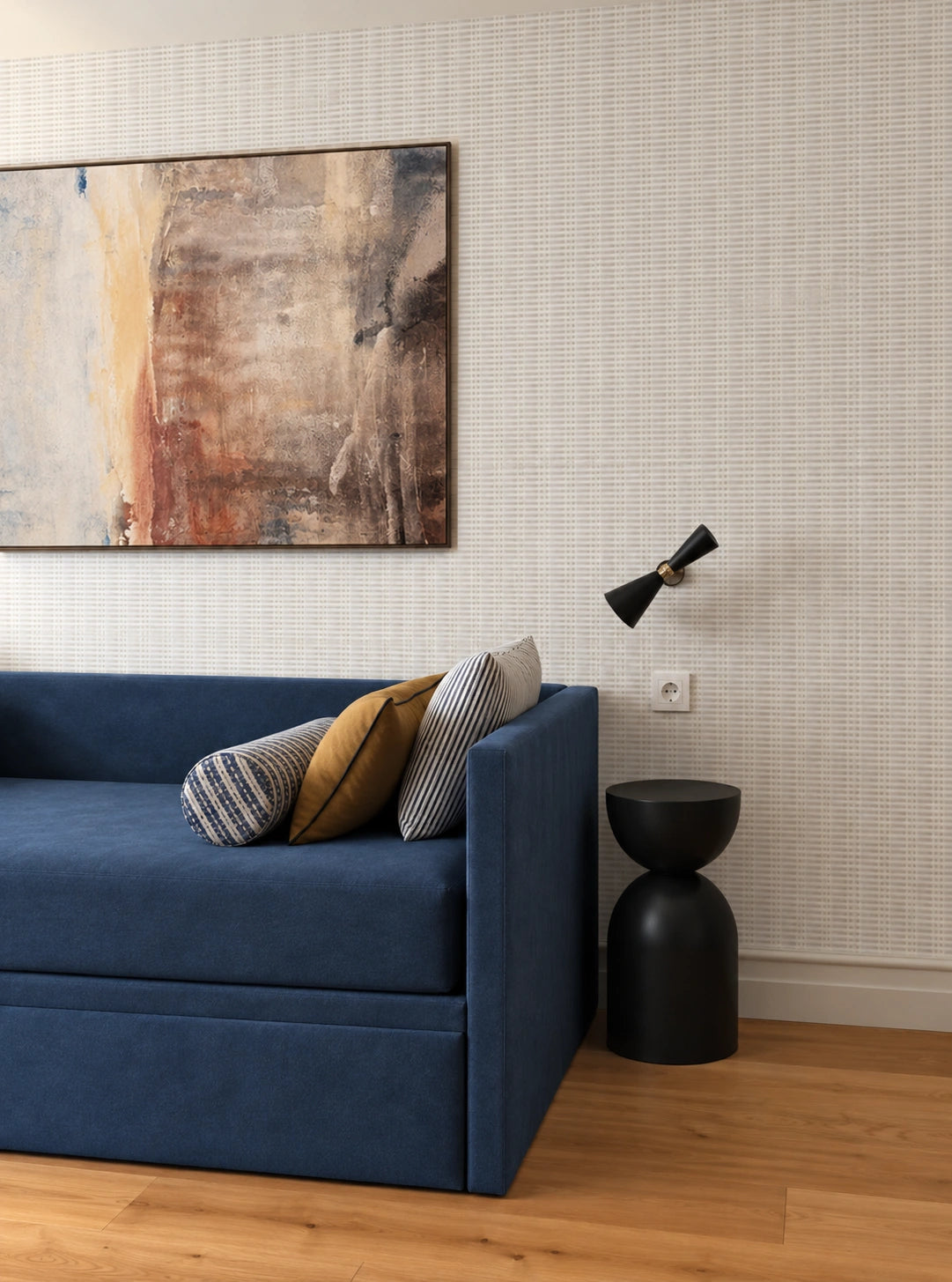 Contemporary living room with a deep blue sofa, striped and mustard accent pillows, textured neutral patterned wallpaper, abstract wall artwork, black sculptural side table, and modern wall lamp over warm wood flooring.
