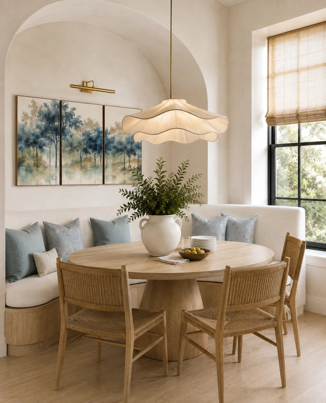 A warm, modern dining nook featuring a round light oak table, four woven-back wooden chairs, a white upholstered banquette with blue pillows, and a large white ceramic vase with green foliage in the center. A sculptural, cloud-like pendant light hangs from the ceiling, and a three-panel landscape painting adorns the arched wall.