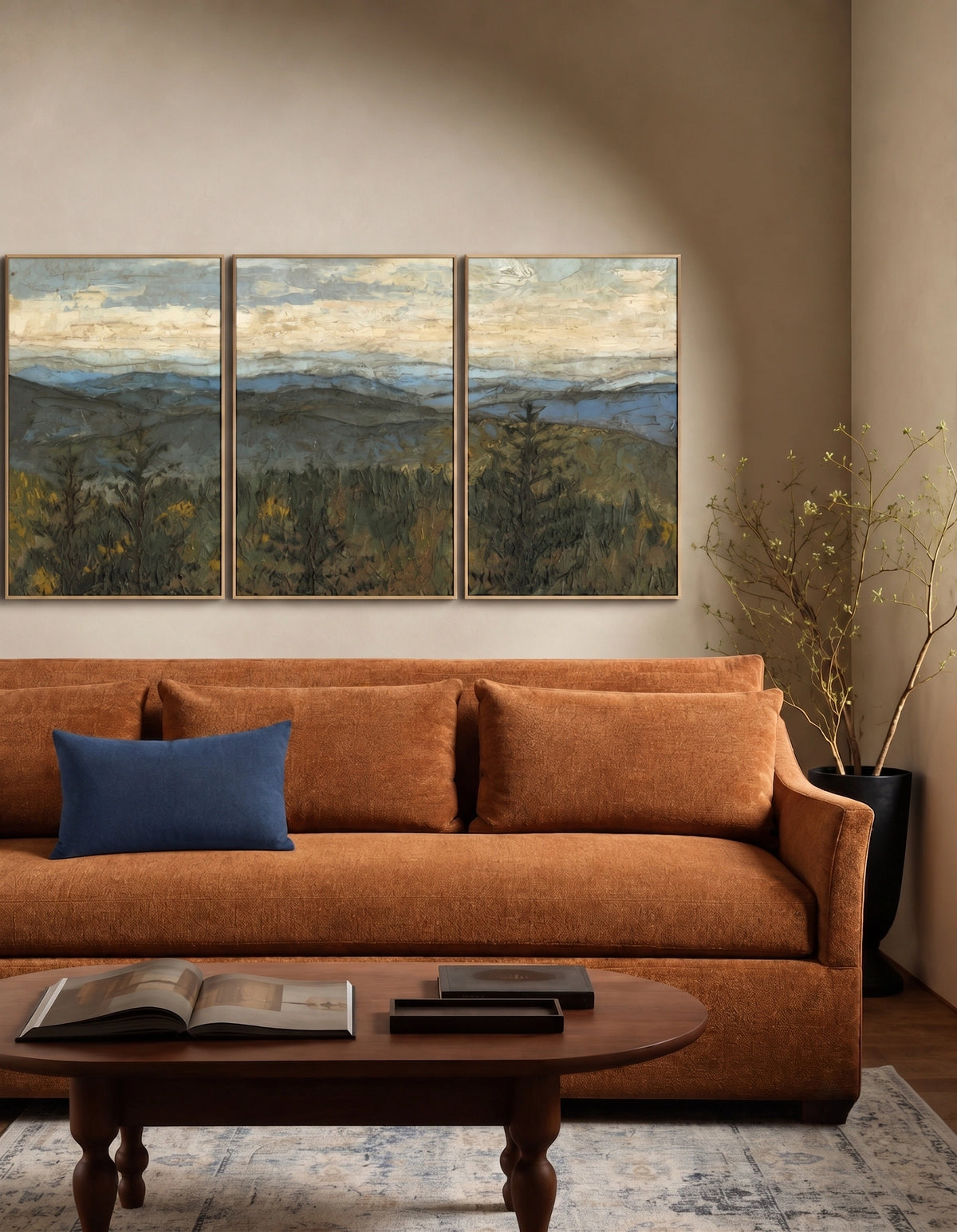 A three-panel framed landscape wall art set hangs above an orange-brown upholstered sofa in a styled living room. The artwork depicts rolling blue mountains and dark green forest trees under a cloudy sky.