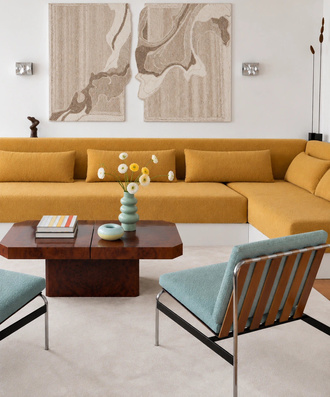Contemporary living room with a mustard yellow sectional sofa, sculptural beige wall art, light blue upholstered lounge chairs, dark wood coffee table, pastel ceramic vases with flowers, and a neutral textured rug.