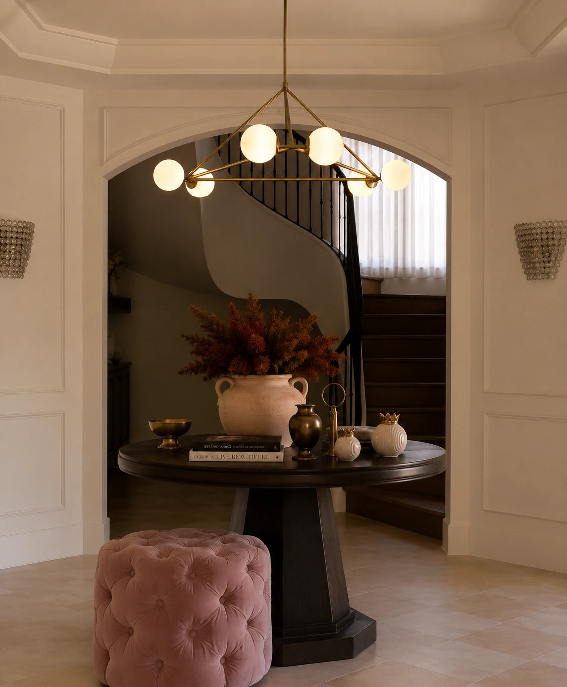A modern brass multi-light chandelier with spherical glass globes hangs over a round dark console table in a decorated entryway. A pink tufted velvet stool sits in the foreground, with a staircase visible in the background.