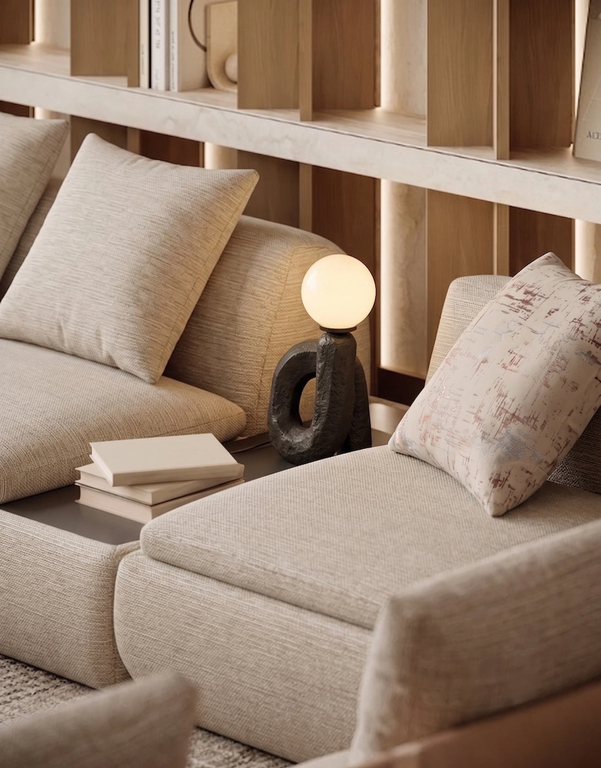 A close-up of a modern beige sectional sofa in a warm, minimalist living room setting, styled with neutral throw pillows, a small black sculptural table lamp with a glowing globe shade, and a stack of books.