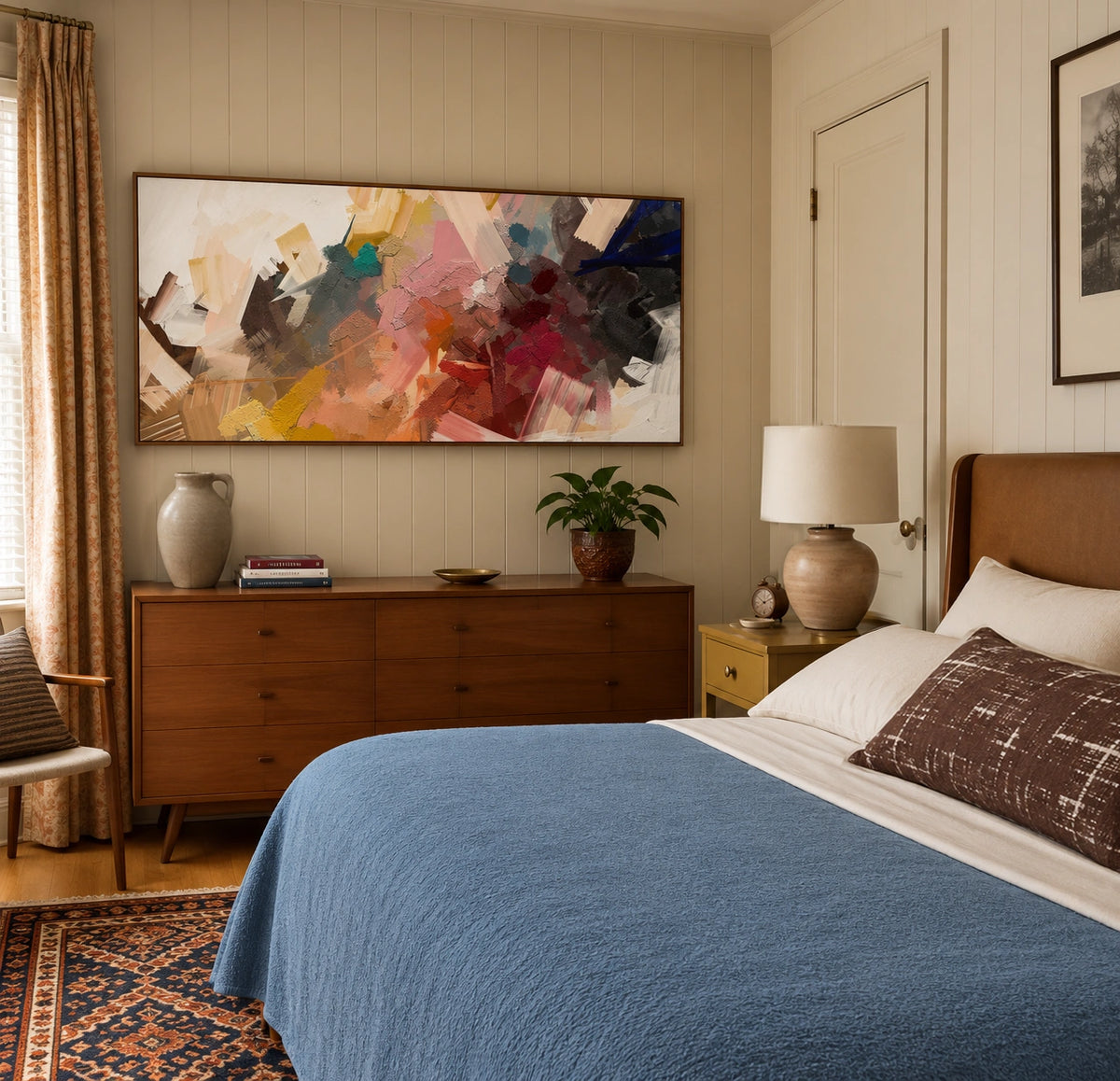 Warm modern bedroom with a blue textured bedspread, brown upholstered headboard, mid-century wood dresser, colorful abstract artwork, patterned area rug, bedside lamp, and neutral paneled walls.