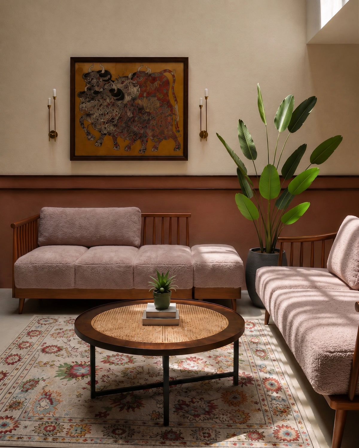 A styled living room interior featuring a mauve-pink upholstered wooden sofa and matching armchair, a round woven coffee table, a patterned area rug, a large potted green plant, and a framed abstract painting on a beige wall.