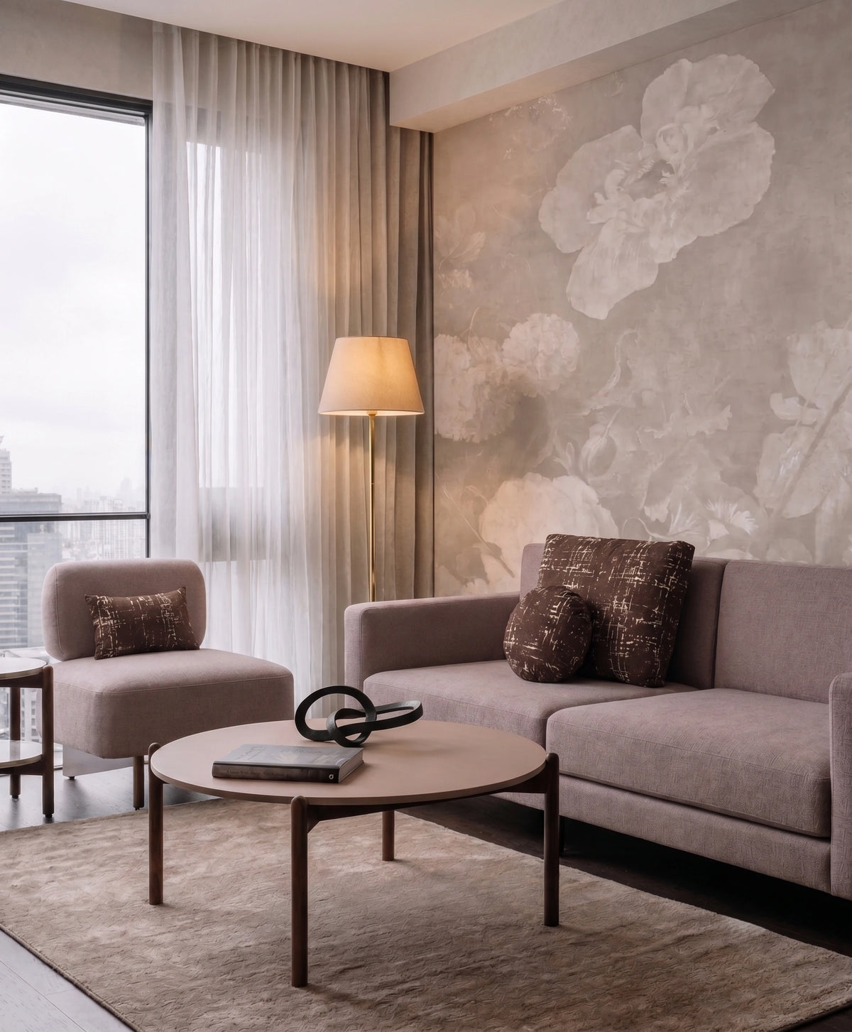 A modern living room featuring a light taupe sofa, a matching armchair, a round coffee table, and a large wall mural with abstract floral patterns in warm beige and gray tones.