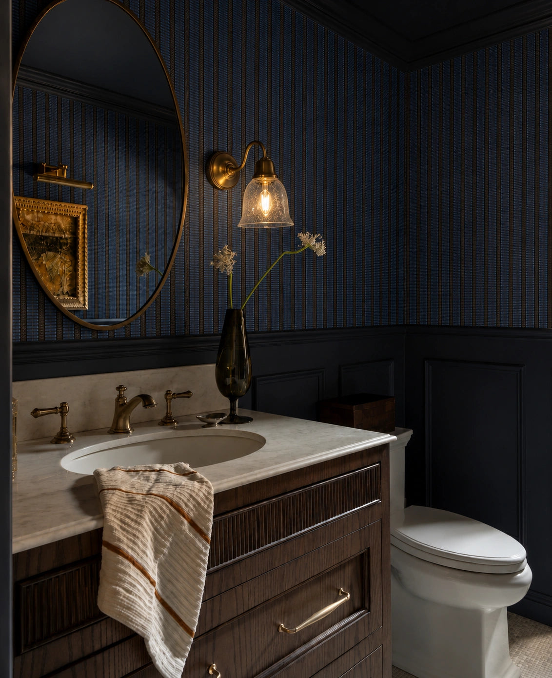 Moody modern bathroom with dark blue textured walls, a wood vanity with marble countertop and brass fixtures, round mirror, warm glass wall sconce, decorative vase, and white toilet.