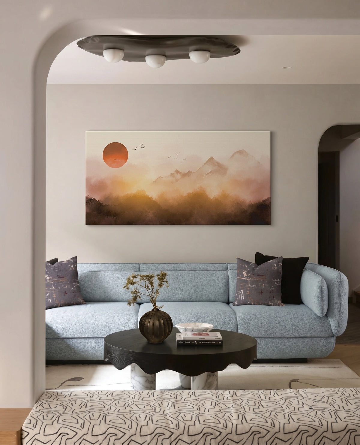 A horizontal landscape canvas print featuring a misty mountain range under a large orange sun, displayed on a light gray wall above a light blue sofa in a modern living room setting.