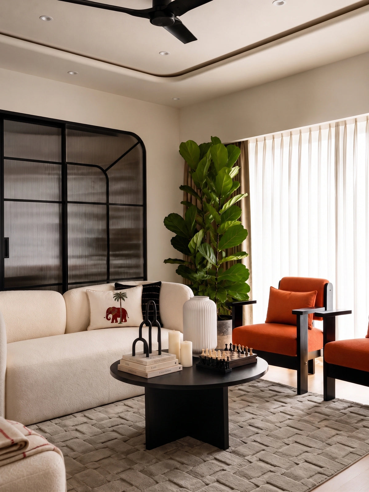 Modern living room with a curved cream sofa, bold orange accent chairs, a round black coffee table, textured neutral rug, indoor fiddle-leaf fig, black-framed glass partition, and soft natural light through sheer curtains.