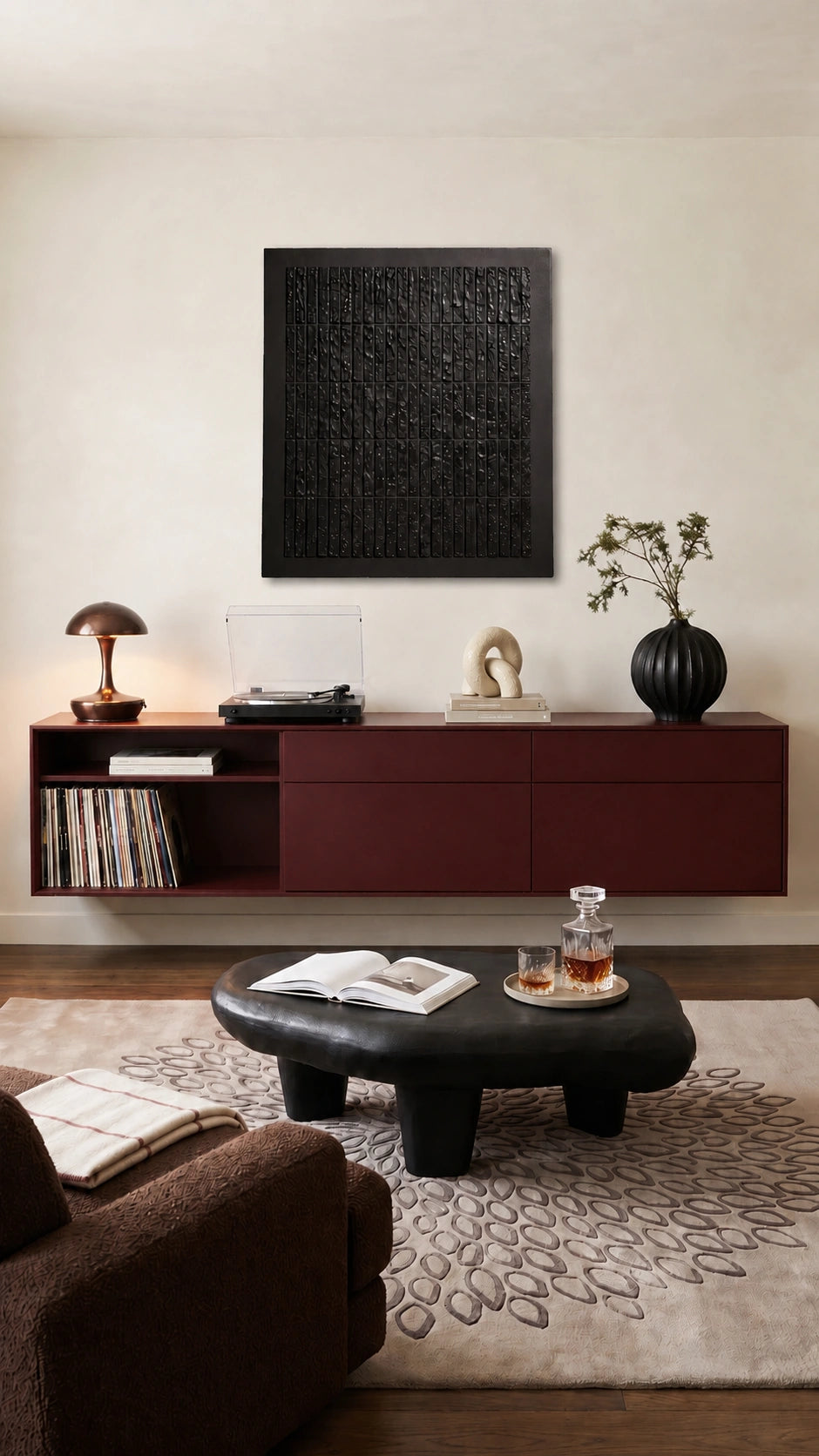A modern living room scene featuring a dark red floating media console against a beige wall, with a black textured square wall art piece mounted above it. The room is styled with a black leather coffee table, a patterned beige rug, and a brown armchair in the foreground.