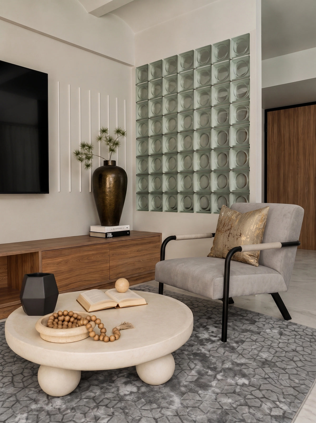 A modern living room featuring a light gray upholstered armchair with black metal arms, a round beige stone coffee table, a gray patterned rug, and a decorative glass block wall in the background.
