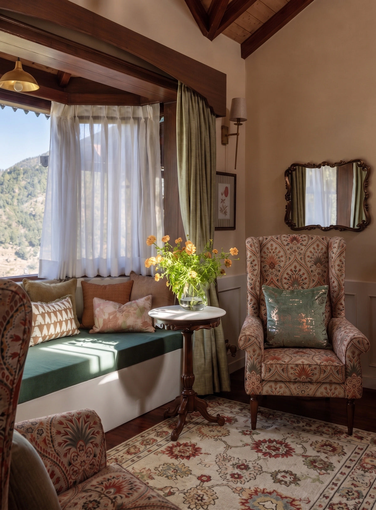 A cozy mountain-view living room corner with a built-in window seat, patterned armchairs, a small round table with orange flowers, and a rug. The room has warm beige walls, dark wood beams, and a window looking out onto a hillside landscape.