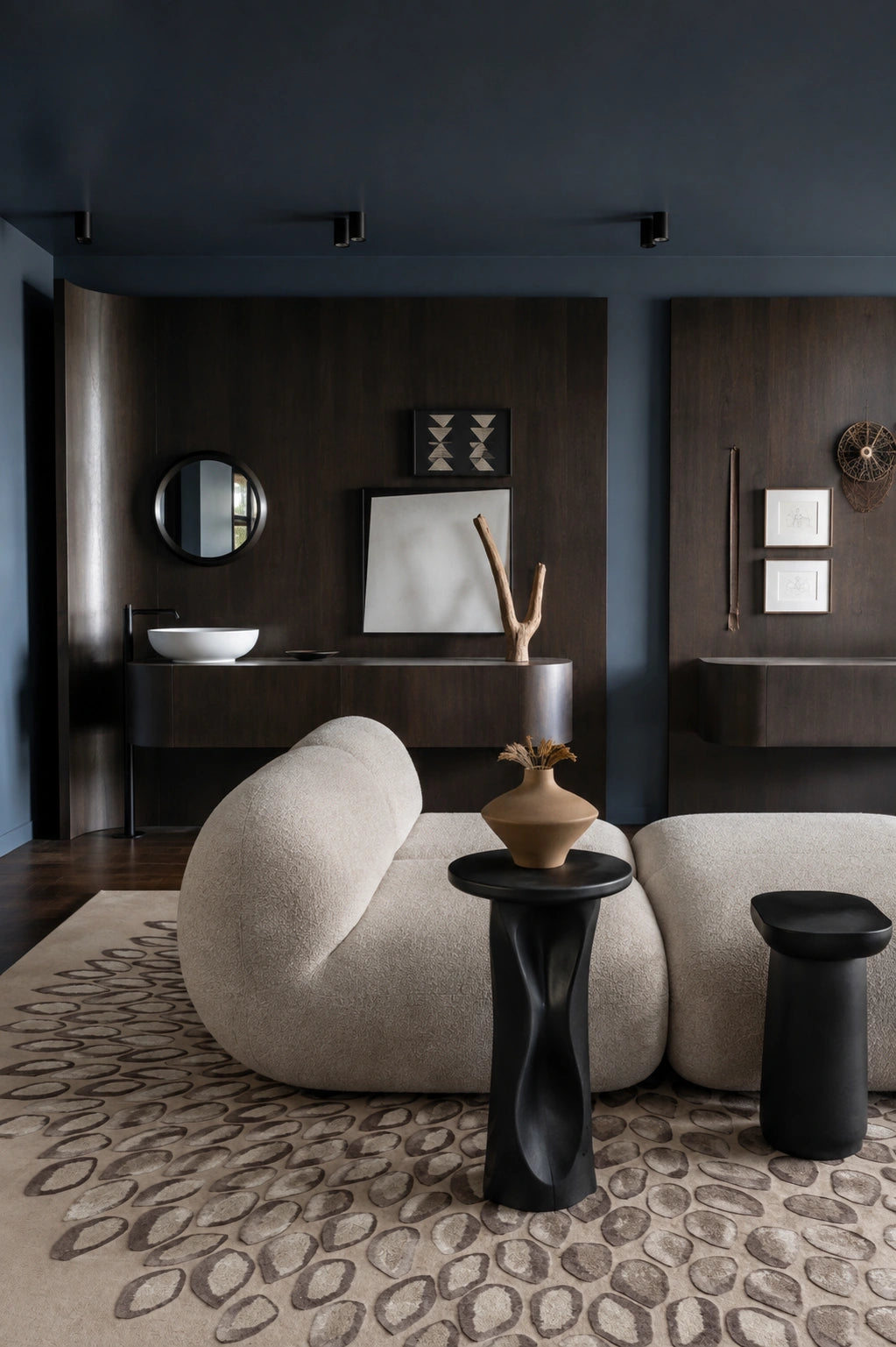 A modern living room interior featuring a curved, low-profile beige bouclé sofa, a black sculptural side table with a ceramic vase, a round black stool, and a patterned beige rug. The background consists of dark wood-paneled walls with a sink, mirror, and framed art.