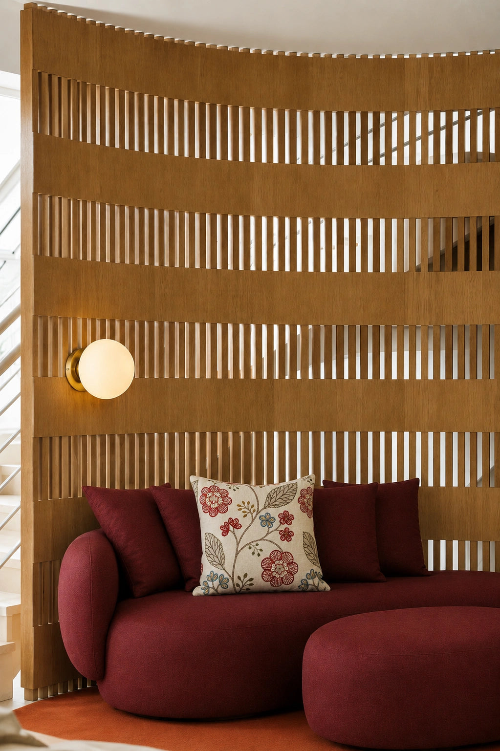 A curved modern sofa upholstered in deep burgundy fabric, styled with matching burgundy pillows and a decorative floral accent pillow, set against a large slatted wooden screen wall with a spherical wall lamp.