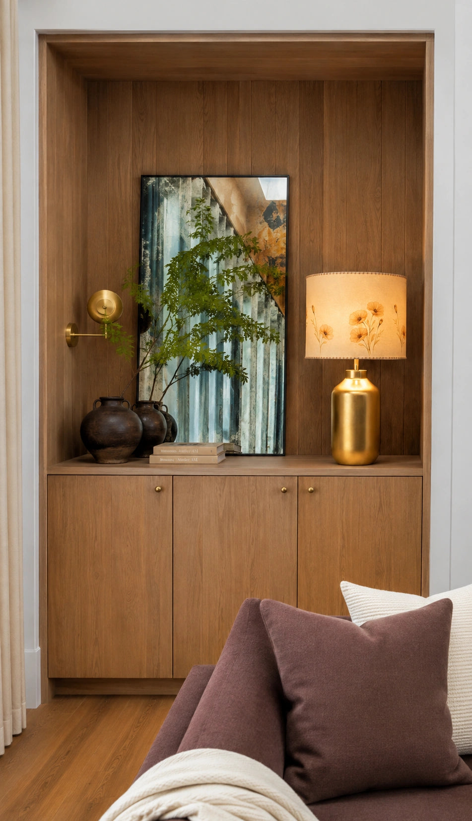 A warm wood cabinet or niche in a living room setting, styled with a gold table lamp, a framed botanical print, dark ceramic vases, and books. In the foreground, a modern sofa with brown and white cushions is partially visible.