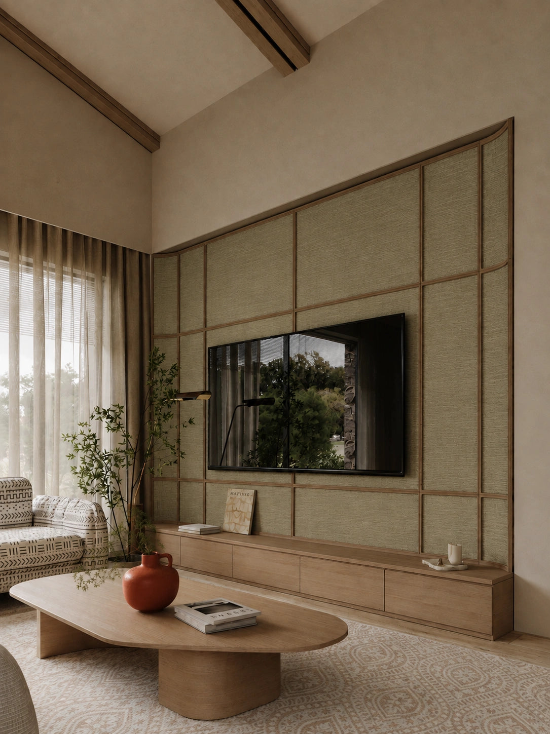 A modern living room interior featuring a large wall-mounted television set within a beige textured panel wall with wooden trim, a low wooden media console, a curved wooden coffee table, and a patterned armchair near a window with sheer curtains.
