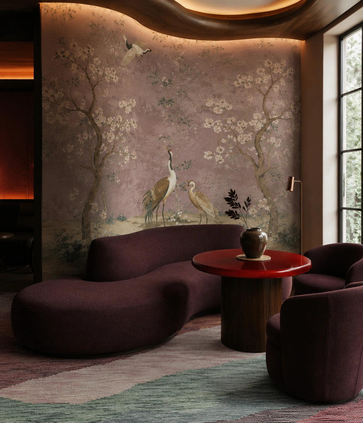 A luxury interior design scene featuring a large wall mural with a traditional Chinese landscape of cranes and cherry blossoms in muted pink and beige tones. In the foreground, there is a modern sculptural curved sofa in deep plum purple, a round red-top wooden coffee table, and a striped area rug.