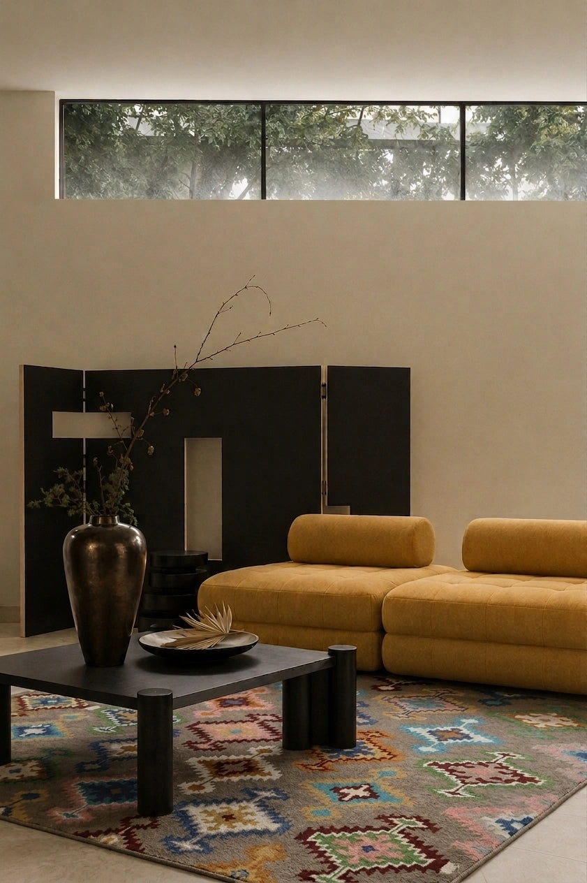 A modern living room scene featuring a mustard yellow modular sofa, a black coffee table with a dark metallic vase, a colorful geometric area rug, and a black folding screen in front of a beige wall with a high window.