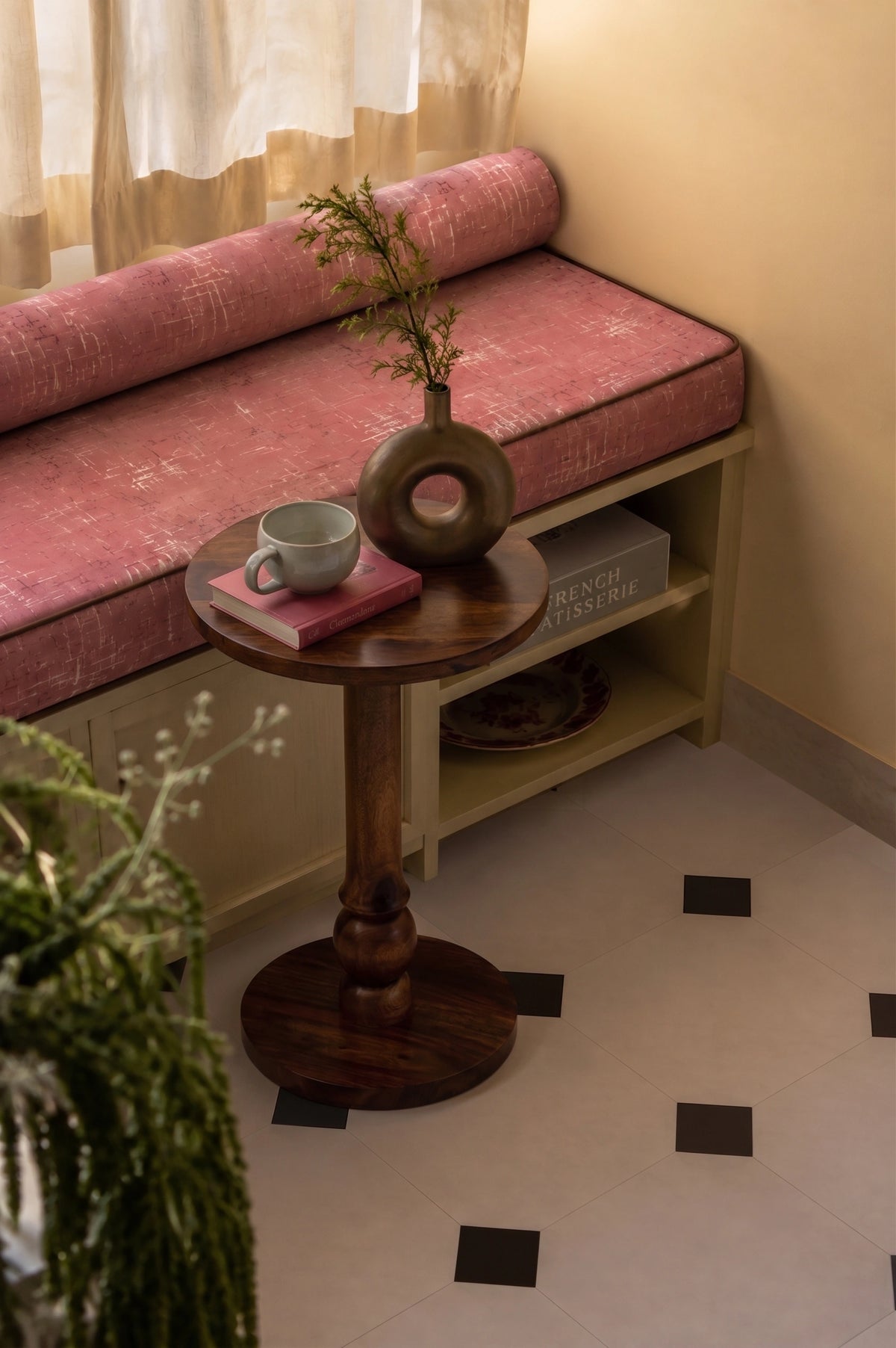 A cozy interior corner featuring a built-in bench with a dusty rose velvet cushion, a round wooden pedestal side table, a dark brown vase with greenery, and a light blue mug on the table. The room has warm beige walls, cream curtains, and a light-colored tiled floor with small black square accents.