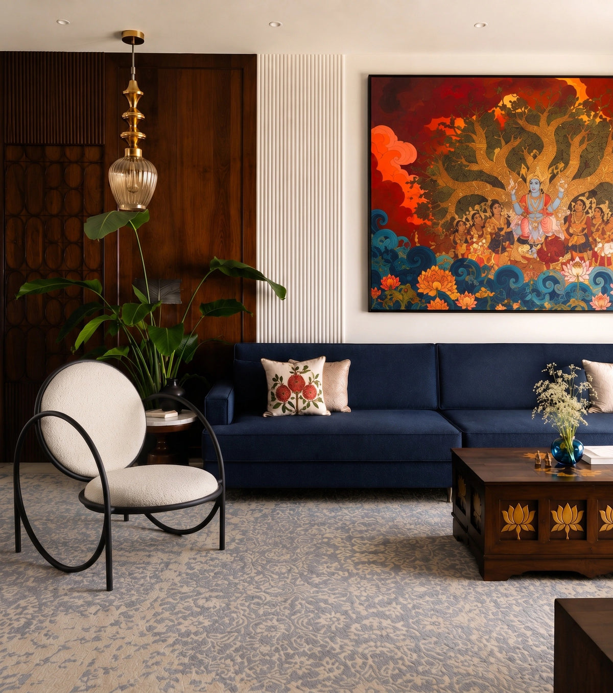A styled living room featuring a large, vibrant traditional-style painting on the wall above a navy blue sofa. The room is decorated with a patterned blue rug, a dark wood coffee table, a white upholstered armchair with a black frame, and a glass pendant light.