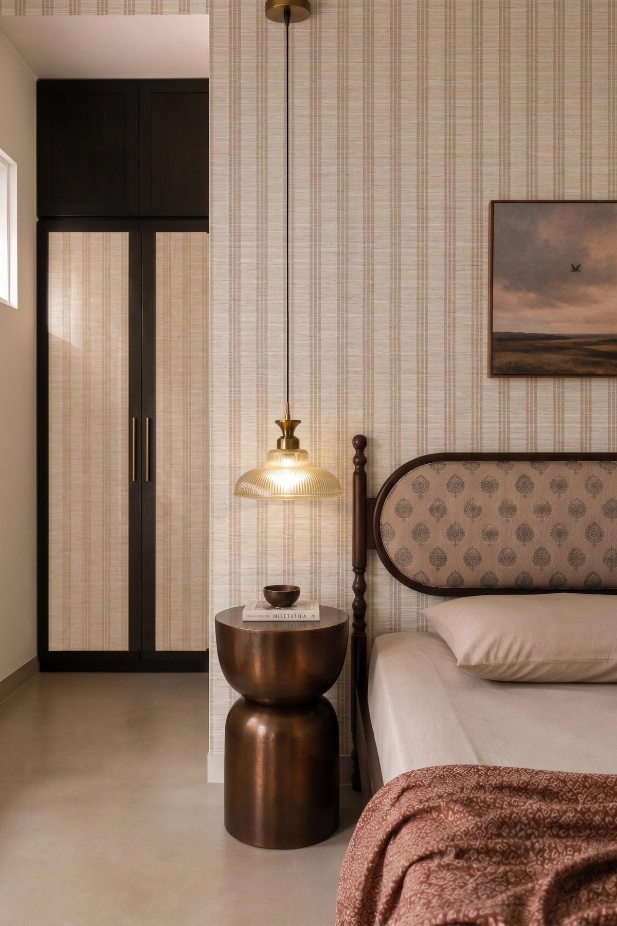 A styled bedroom interior featuring a bed with a patterned headboard, a dark wood nightstand with a glowing pendant lamp, striped wallpaper, and a dark wood wardrobe in the background.