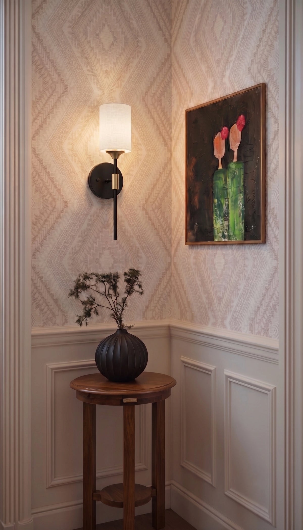 A styled interior corner featuring textured wallpaper, a black wall sconce with a white shade, a framed painting, and a small wooden side table holding a dark vase with dried branches.