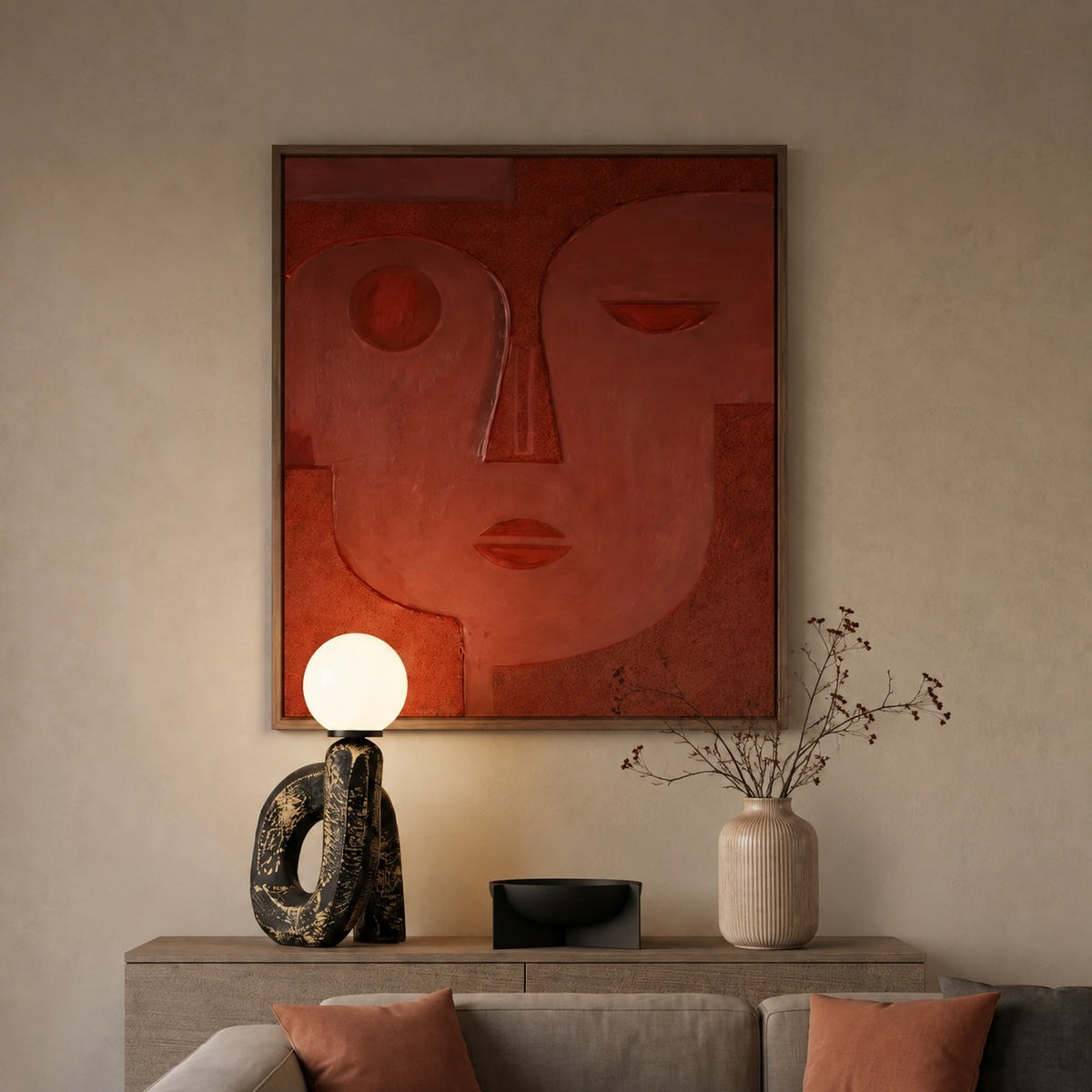 A framed abstract wall art piece featuring a stylized red and terracotta face, displayed above a console table in a modern living room setting with a lamp, vase, and sofa.
