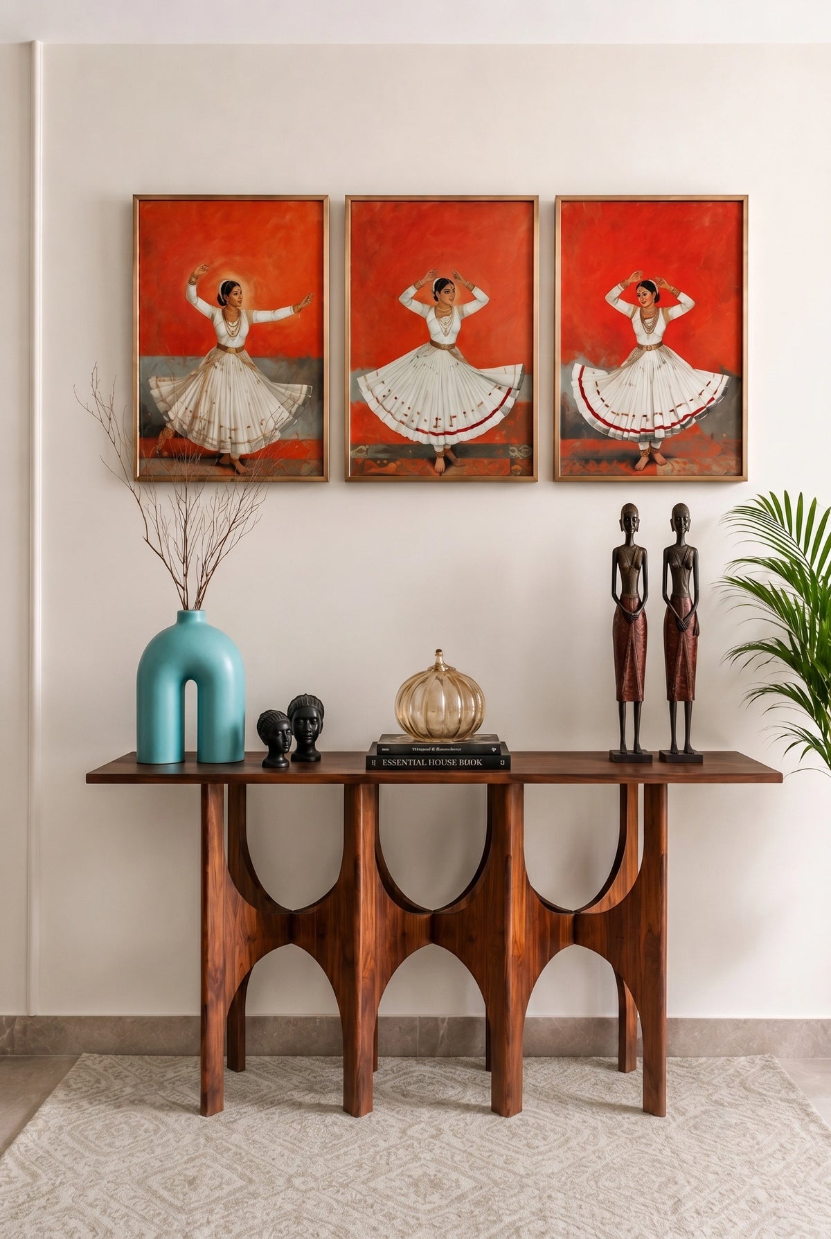 A styled interior scene showing a dark wood console table against a beige wall, decorated with a turquoise vase, dried branches, sculptural objects, and two tall human figurines. Above the table are three framed paintings of dancers against vivid red backgrounds.