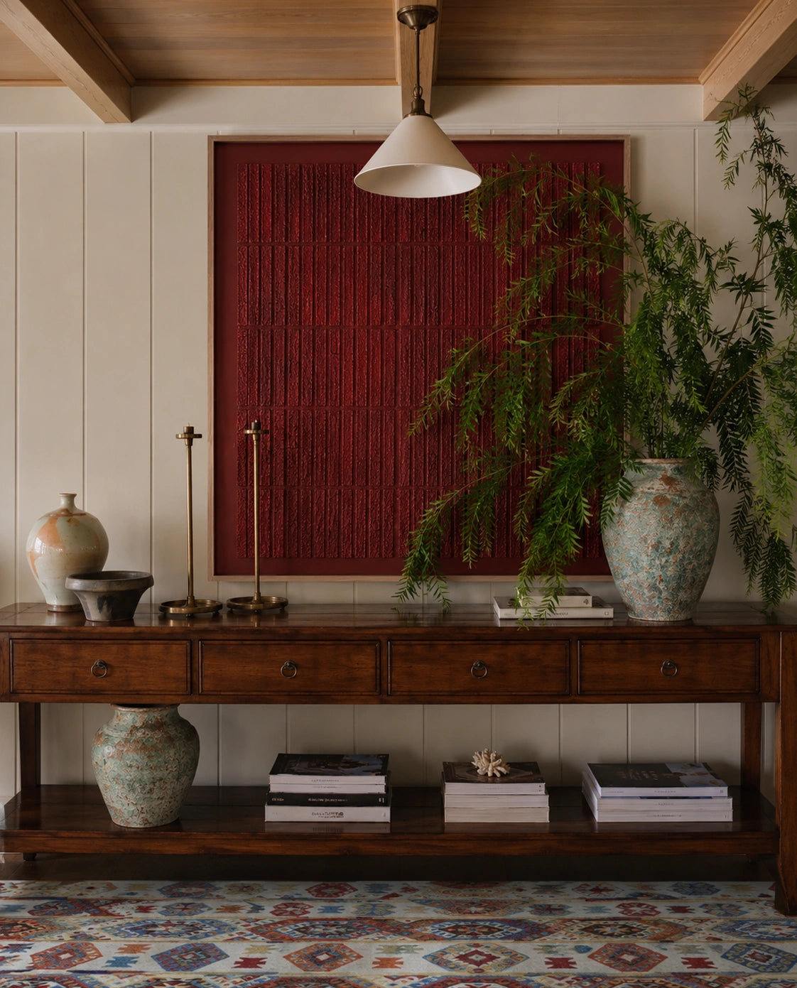 A styled interior scene featuring a large, square, deep red wall art piece with vertical slats, mounted on a cream paneled wall above a dark wood console table. The room is decorated with a hanging pendant light, a tall green fern in a patterned ceramic vase, and other decorative vases and books.