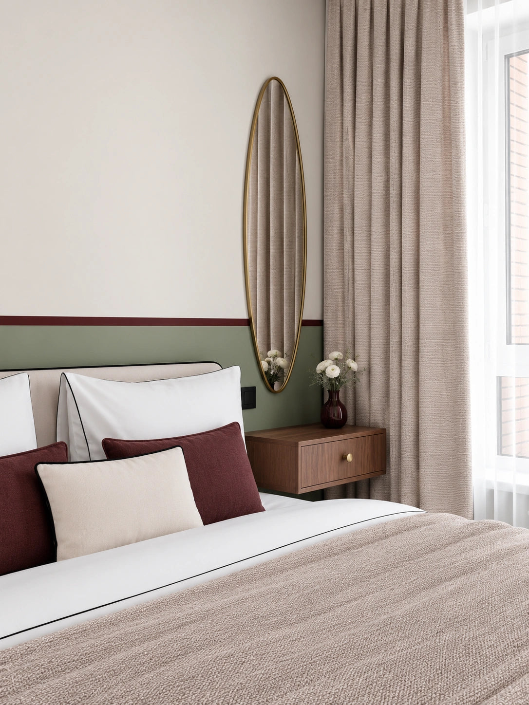 Elegant bedroom with a white upholstered bed, burgundy and cream accent pillows, sage green and cream paneled walls, a slim gold oval mirror, wood bedside table, floral vase, and textured beige curtains beside a bright window.