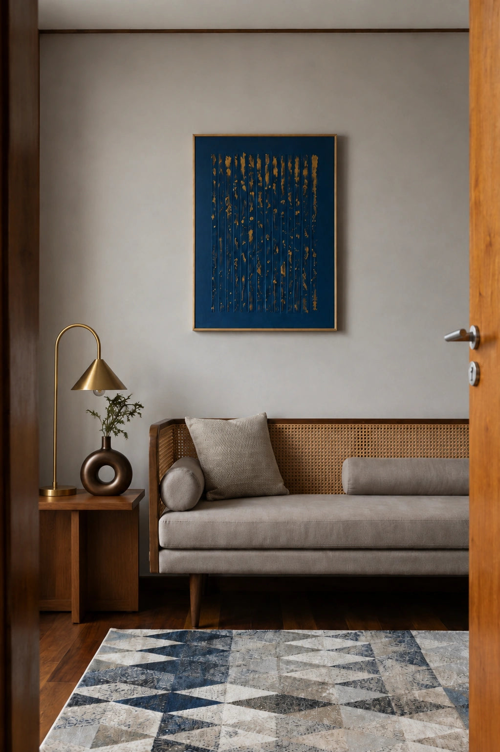 A modern living room scene featuring a gray fabric sofa with a woven cane backrest, a brass floor lamp on a wooden side table, and a framed abstract blue and gold wall art piece above the sofa.