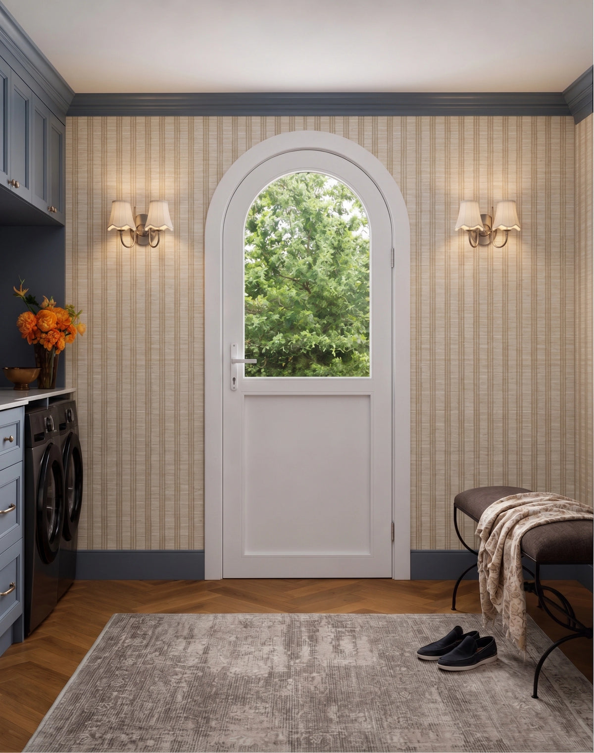 A white arched entryway door with a glass window at the top, set in a tiled laundry room with beige striped wallpaper, wood floors, a gray rug, and a bench with a throw blanket.
