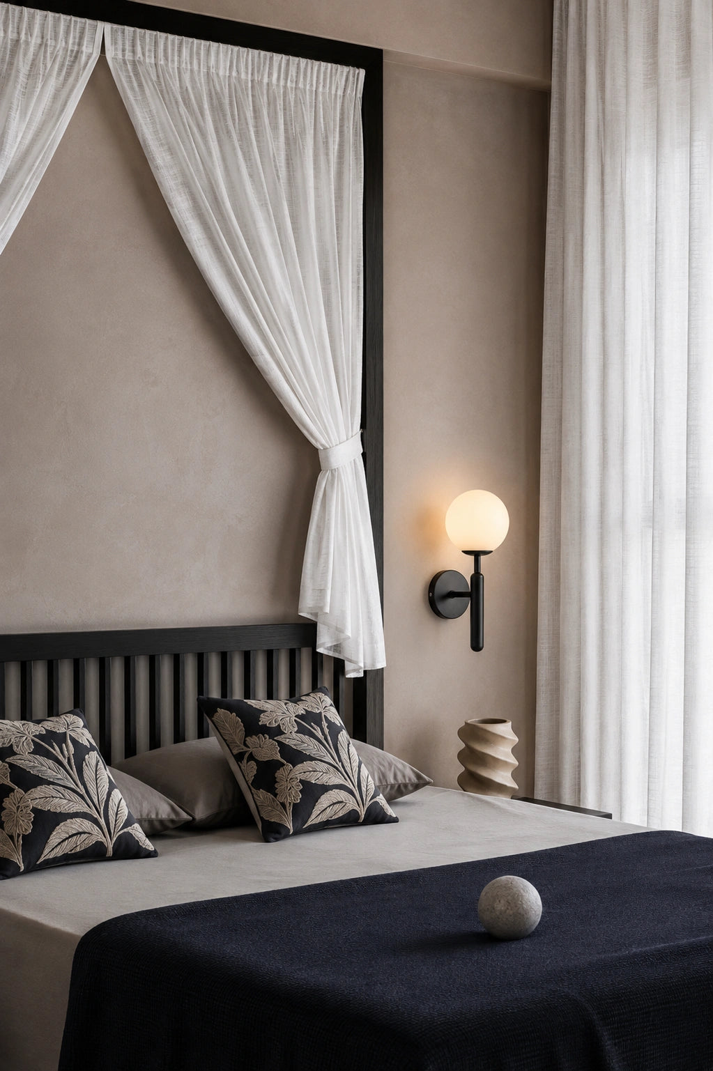 A modern bedroom scene featuring a bed with a dark wooden slatted headboard, white draped curtains, patterned pillows, and a dark navy blue bedspread. A black wall-mounted spherical lamp is lit on the beige plaster wall next to the window.