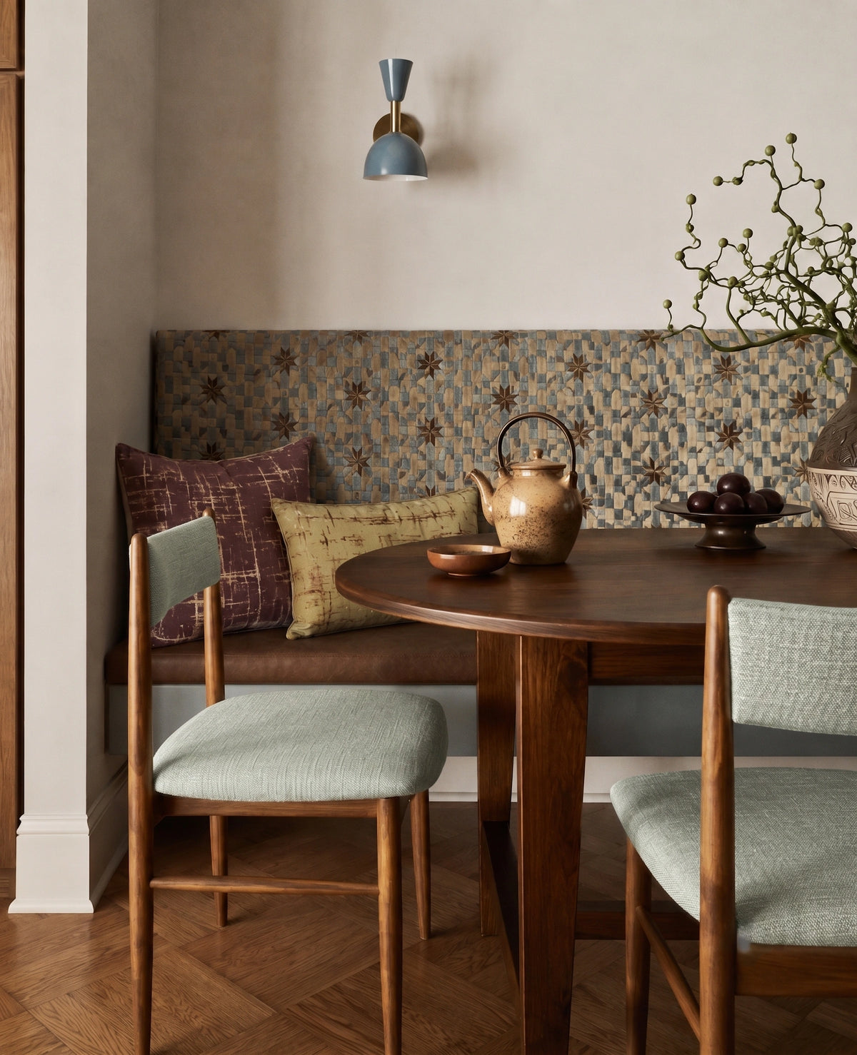 A cozy dining nook with a round wooden table, two upholstered chairs, a patterned bench, and decorative pillows in a warm neutral interior.