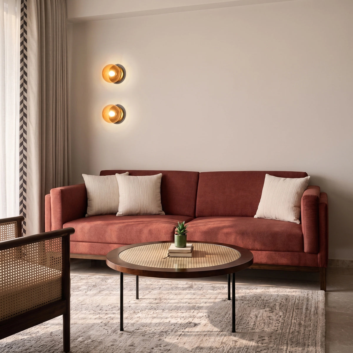 A modern living room scene featuring a rust-red velvet three-seater sofa with two cream throw pillows, set against a warm beige wall with two glowing circular wall sconces. A round coffee table with a woven cane top sits in front of the sofa on a light gray patterned rug.