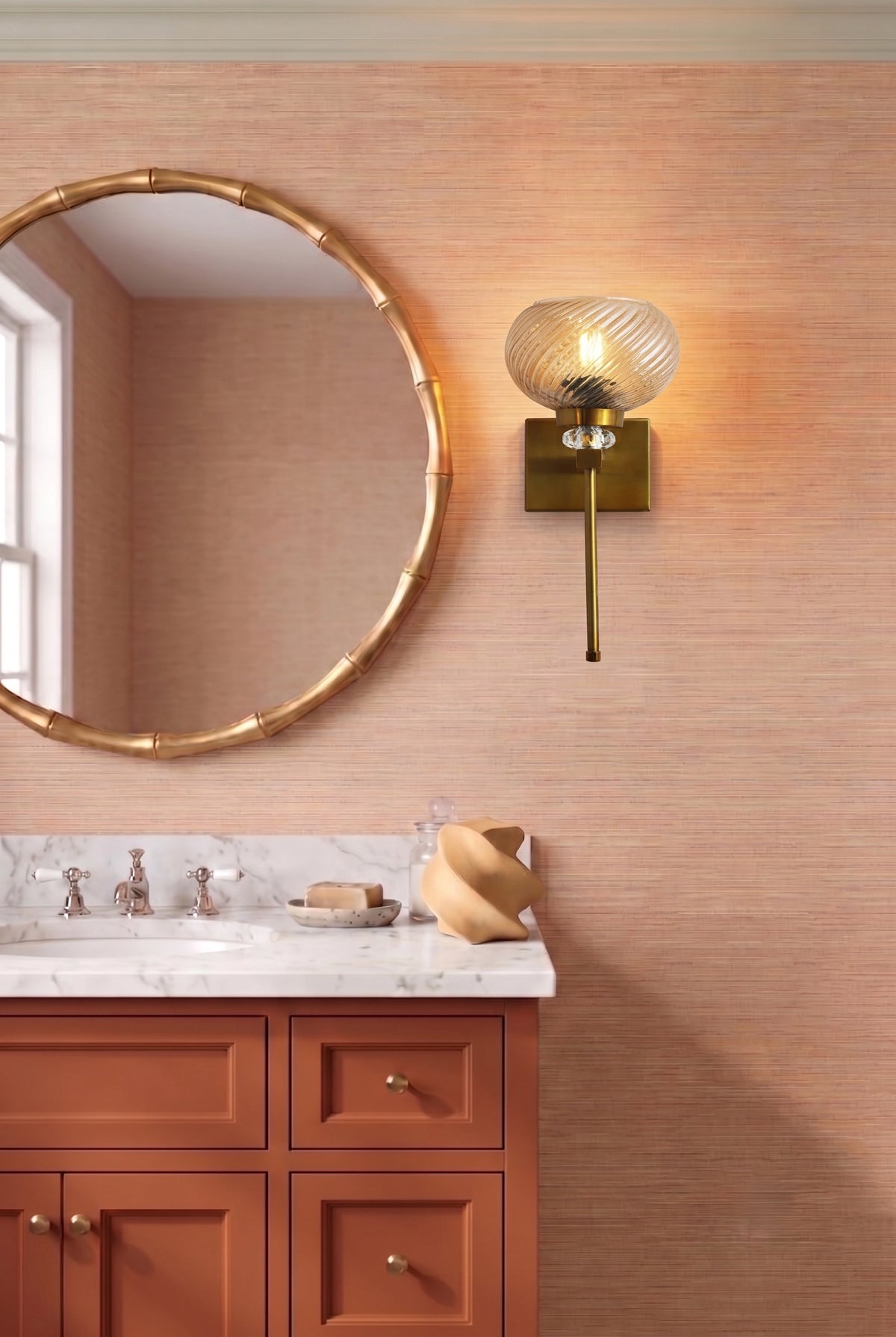 A warm-toned bathroom interior featuring a round gold-framed mirror, a wall-mounted brass sconce with a ribbed glass shade, a terracotta vanity cabinet, and a white marble countertop.