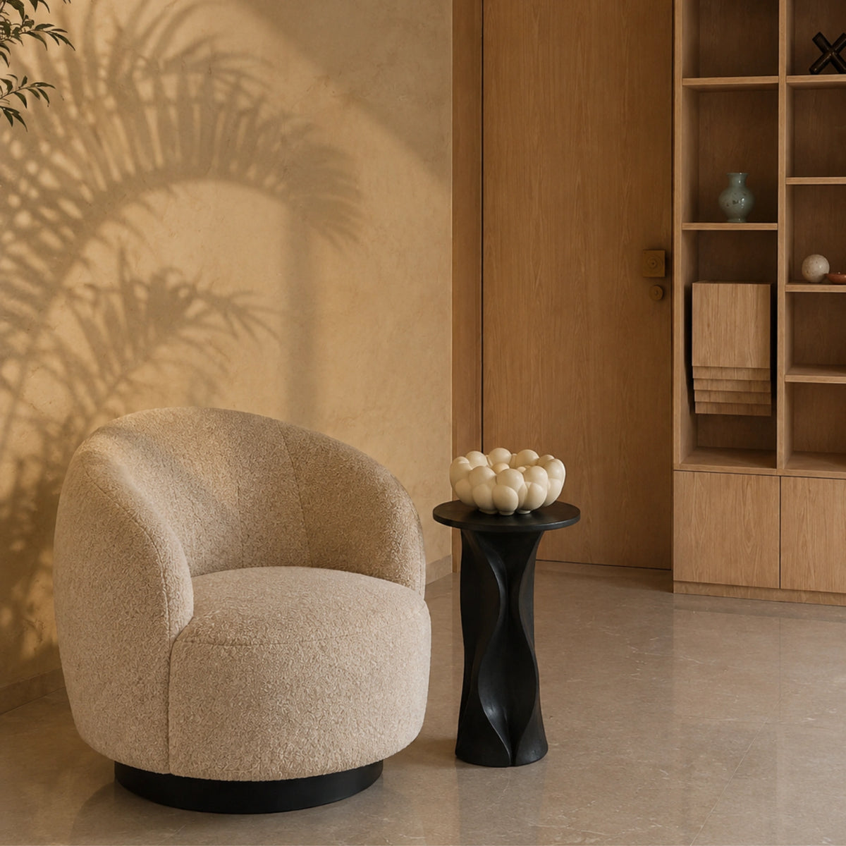 A modern round bouclé armchair in a warm beige color sits next to a sculptural black side table in a minimalist interior setting with a wooden door and shelving in the background.