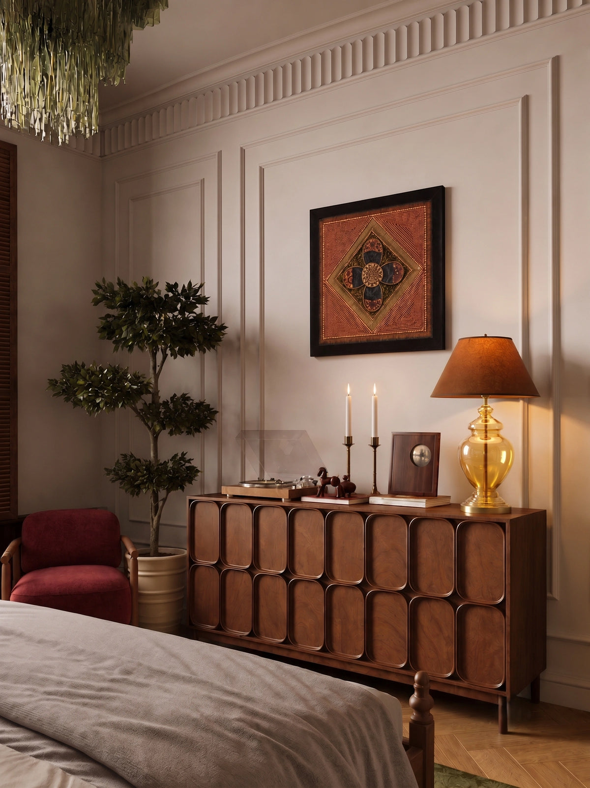 A warm, elegantly styled bedroom corner featuring a mid-century modern wooden sideboard with a gold lamp, a red armchair, a potted tree, and a framed geometric tapestry on a paneled cream wall.