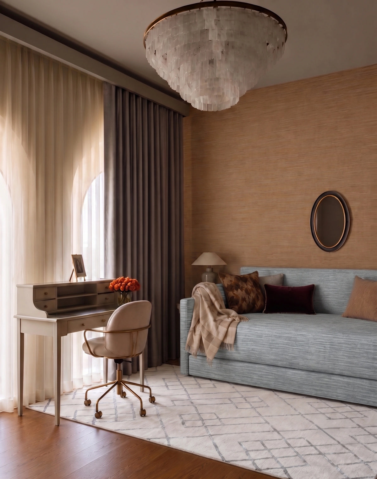 Elegant bedroom with a gray upholstered daybed, neutral and burgundy accent pillows, wood-textured feature wall, round mirror, vintage writing desk and chair, sheer curtains, dark drapes, patterned area rug, and a statement crystal chandelier.