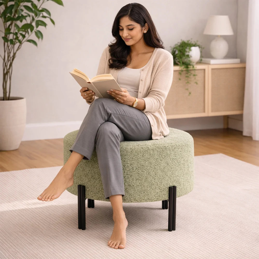 A woman sits barefoot on a round, textured light green upholstered ottoman with black legs, reading a book in a bright, modern living room.
